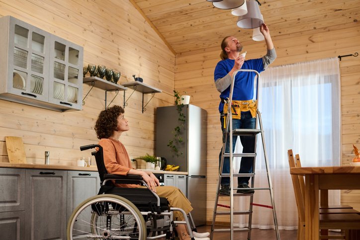 Young housewife with disability sitting in wheelchair in the middle of kitchen and looking at electrician changing bulbs in chandelier